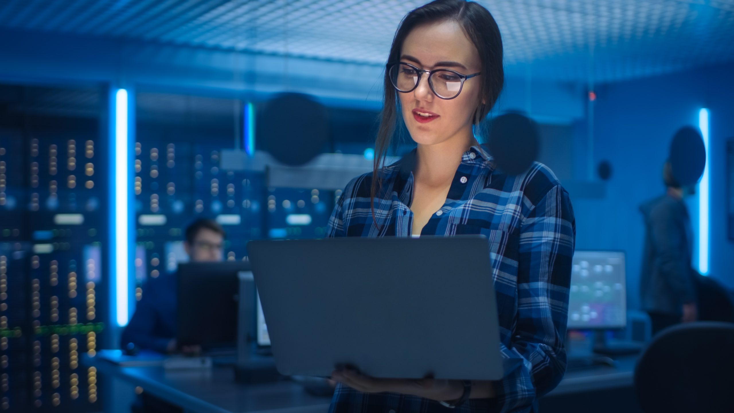 Portrait of a Smart Young Woman Wearing Glasses Holds Laptop. In the Background Technical Department Office with Specialists Working and Functional Data Server Racks