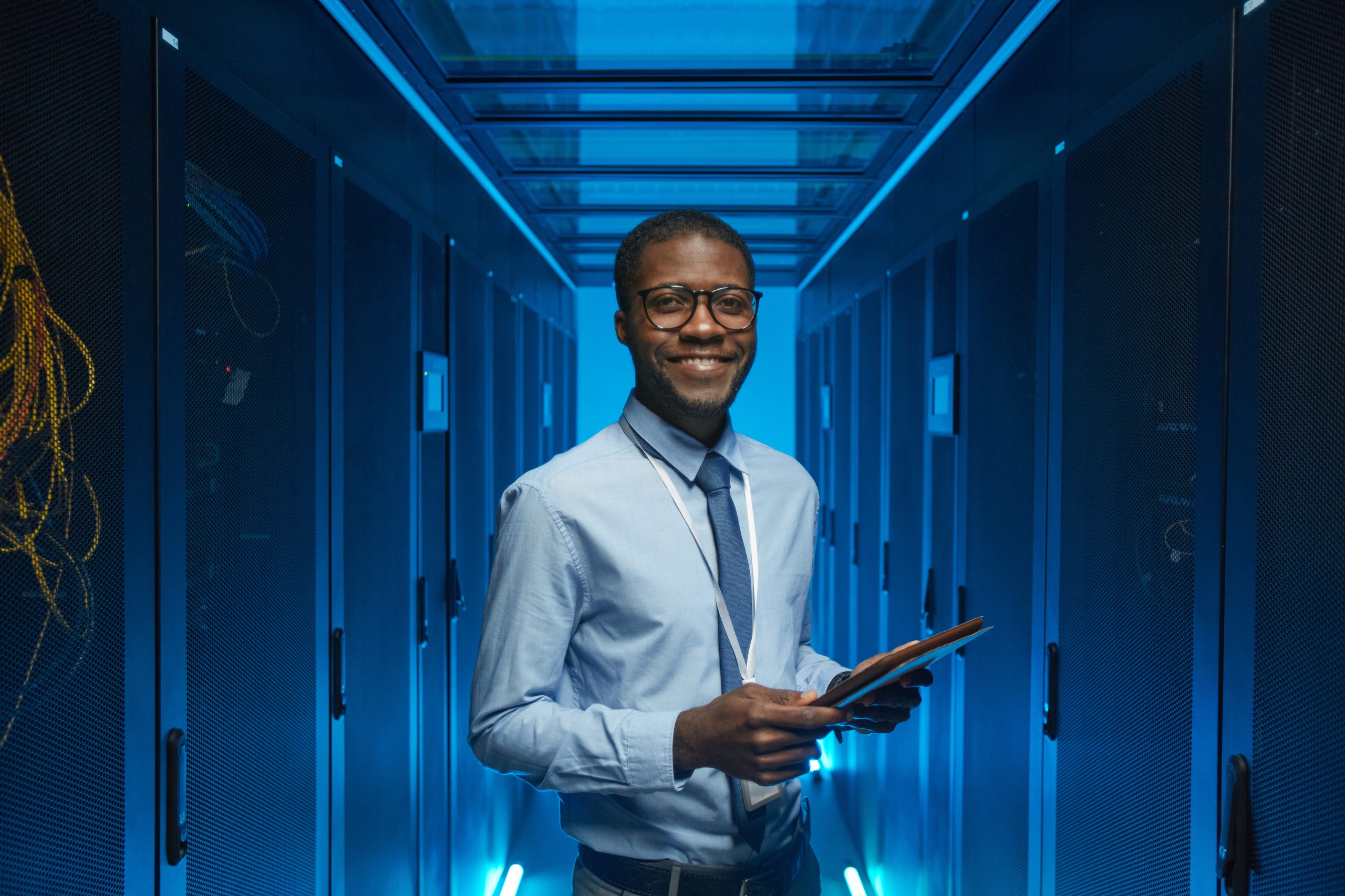 Waist up portrait of smiling man standing by server cabinet while working with supercomputer in data center and holding tablet, copy space